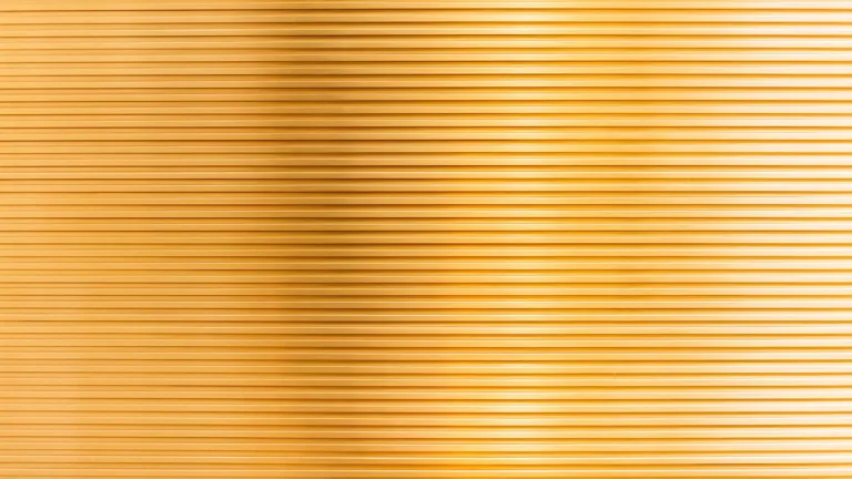 A close-up textured photo of a gold material. The material has small, repeated, horizontal groves creating long lines that cover it entirely.