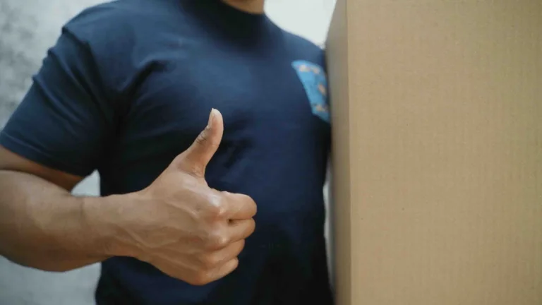 A video of a man holding a brown cardboard box. We can only we the man's torso and the box. Everything else is out of frame. The man is holding up a thumbs up with his other hand. The camera pans from left to right. The man is wearing a navy polo.